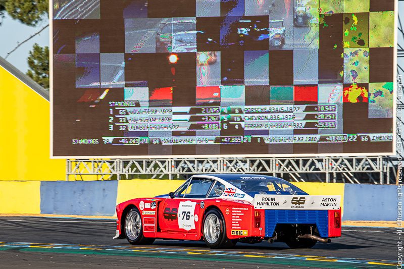 Aston Martin RHAM/1 rouge, blanche et bleue n° 76 vue de trois quarts arrière devant un écran géant au Le Mans Classic 2022.